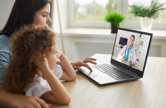 A woman and child sitting at a table with a laptop, engaging with a doctor on the screen.